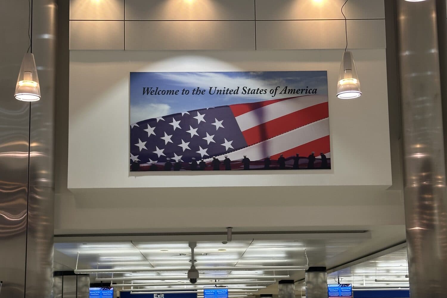 A “Welcome to the United States of America” sign above the arrivals hall, passengers walking toward the exit beneath it