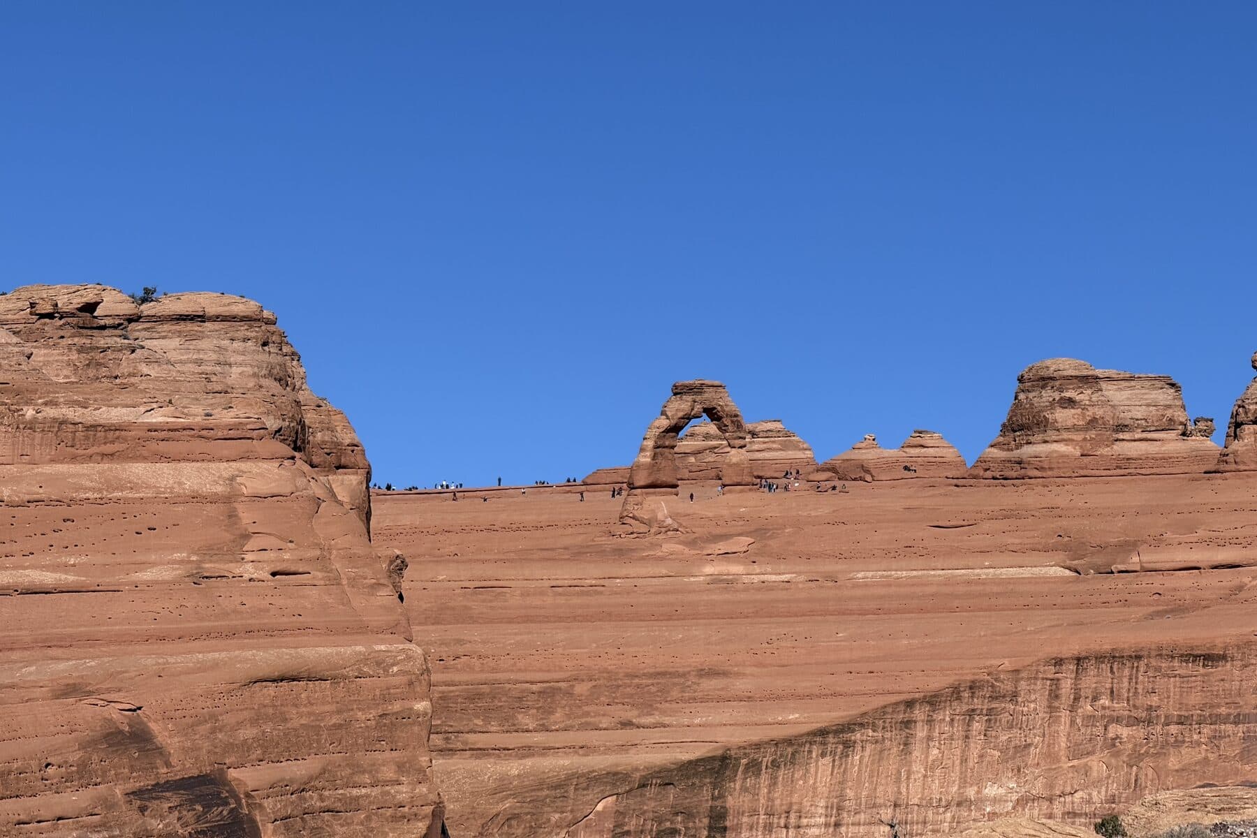 Delicate Arch standing on the far rim of the sandstone bowl, the walkers along the ridge no taller than a fingernail, Arches National Park, Utah