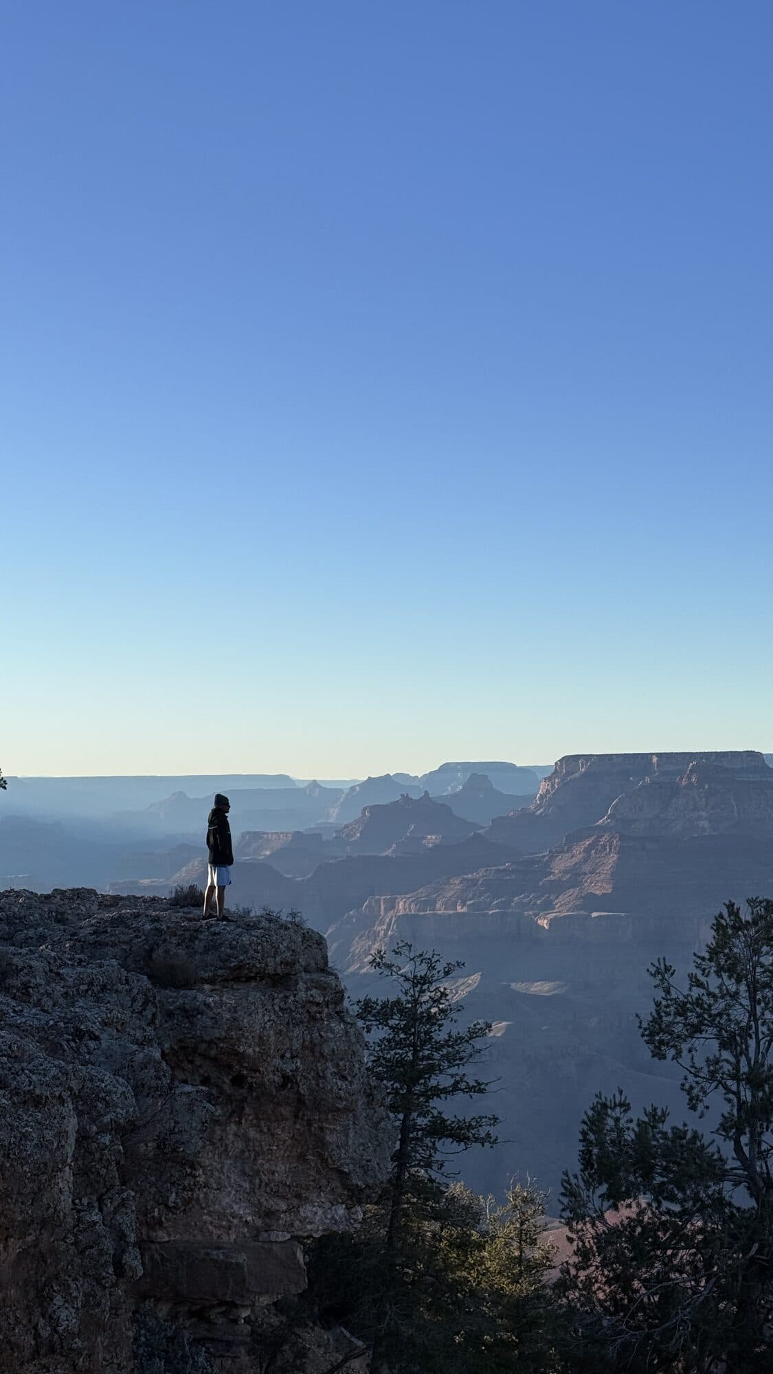 A lone figure standing on a rock ledge at the rim of the Grand Canyon at first light, the canyon walls receding into blue haze behind