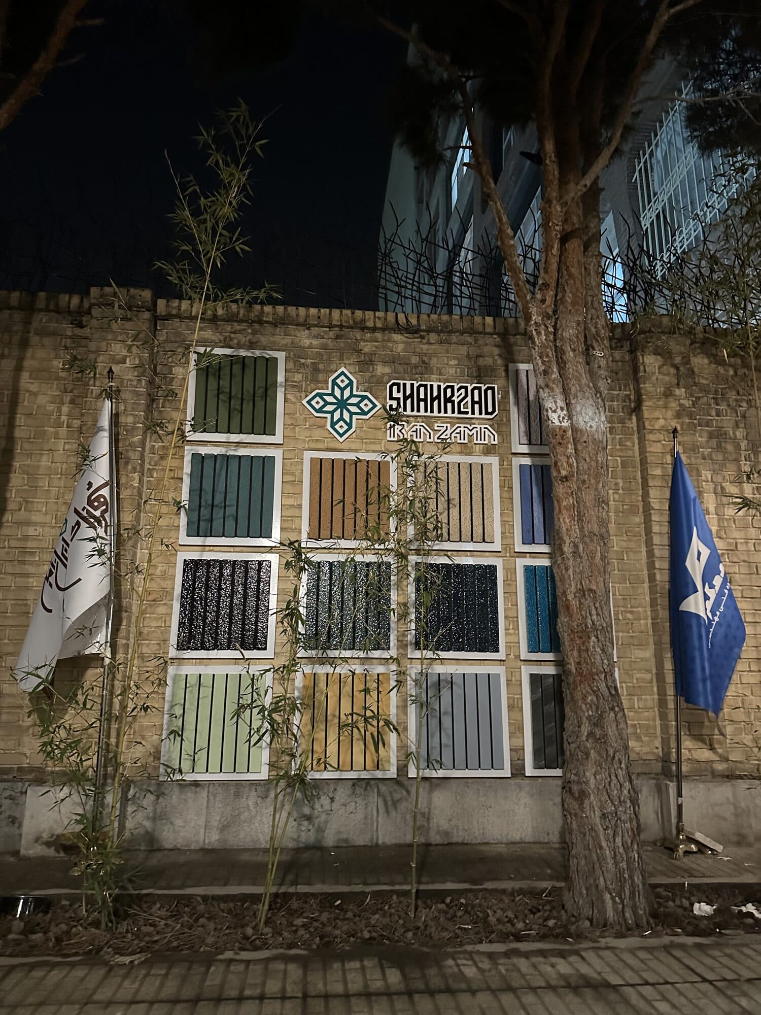 The club from the street at night — a wall of colour-sample panels lit behind young trees, the SHAHRZAD mark above them