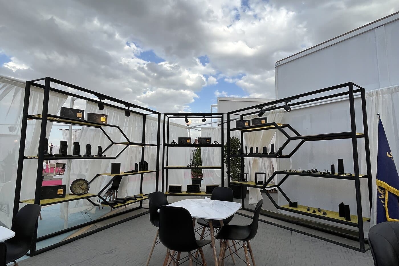 An open black steel grid hung with product and set with a white table under a broken sky — WINDOW SCENE, Mashhad 2023