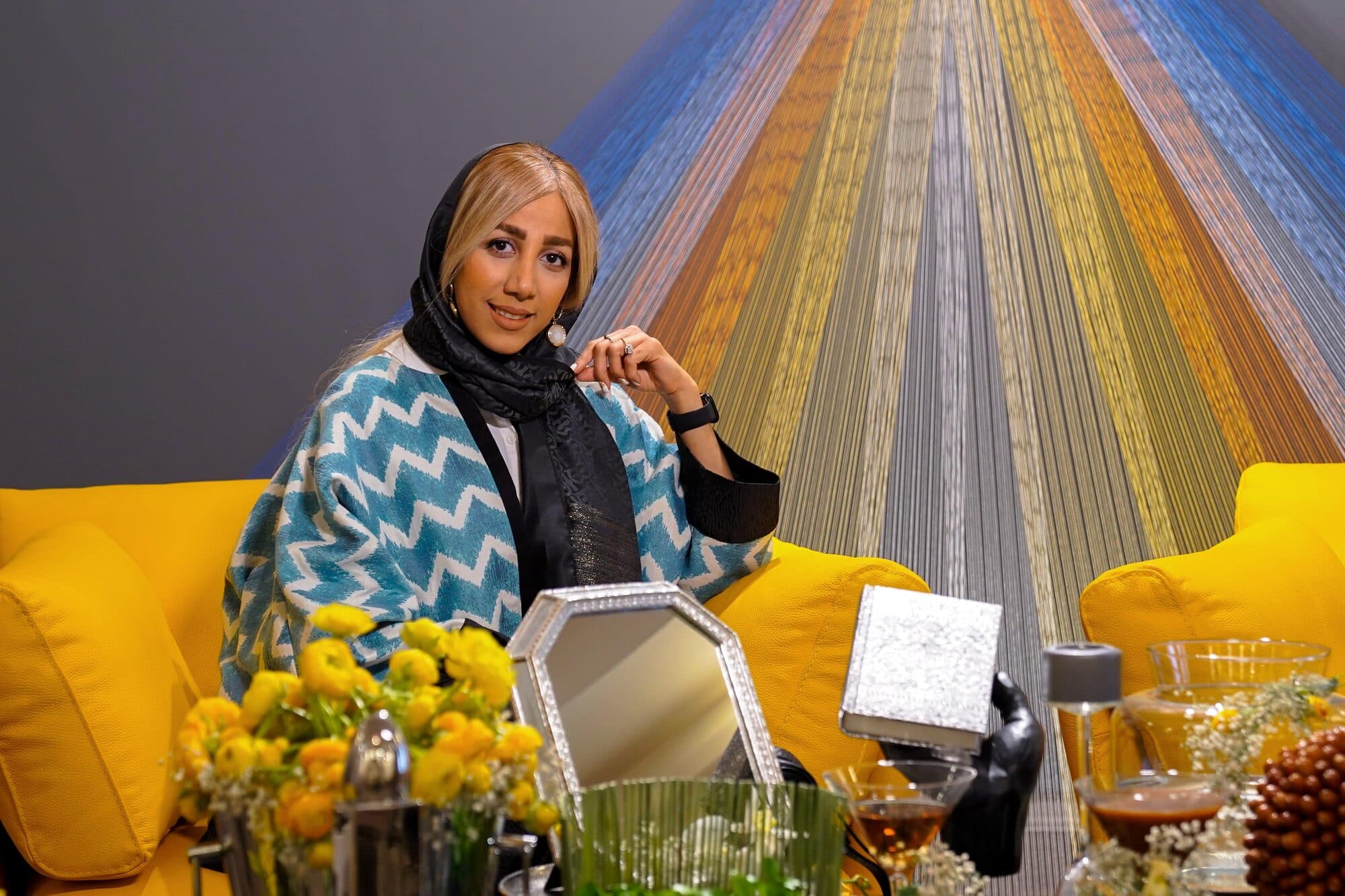 مهمانی با پالتوی طرح‌دار، نشسته در گوشه‌ی دوربین و تکیه‌داده به دسته‌ی مبل.