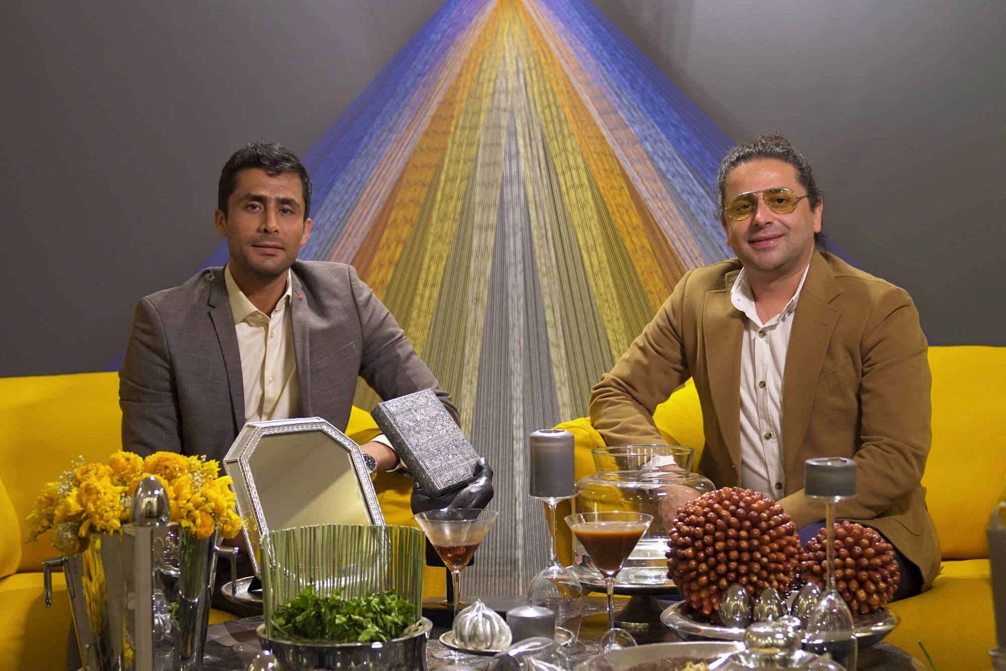 Two guests seated on the yellow sofa in front of the woven thread backdrop, the Haft-Seen laid out between them and the camera.