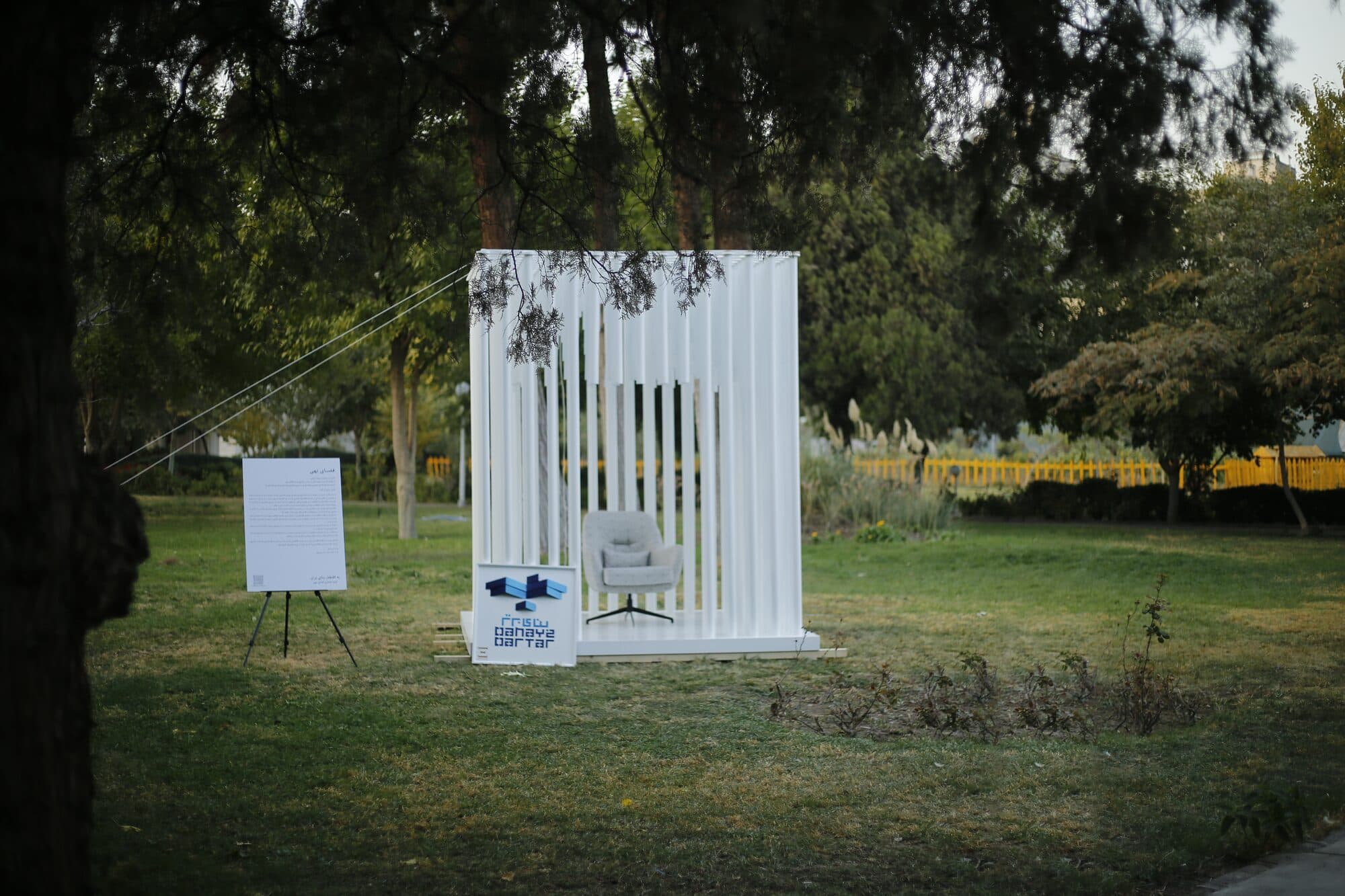 A white-slatted pavilion standing on open lawn under the park's trees, a single armchair inside it and the Banaye Bartar mark on the panel alongside