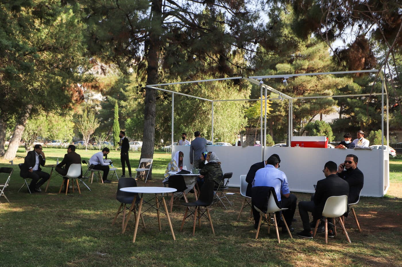 Groups in conversation at open-air lounge settings under the trees, the pavilion behind them