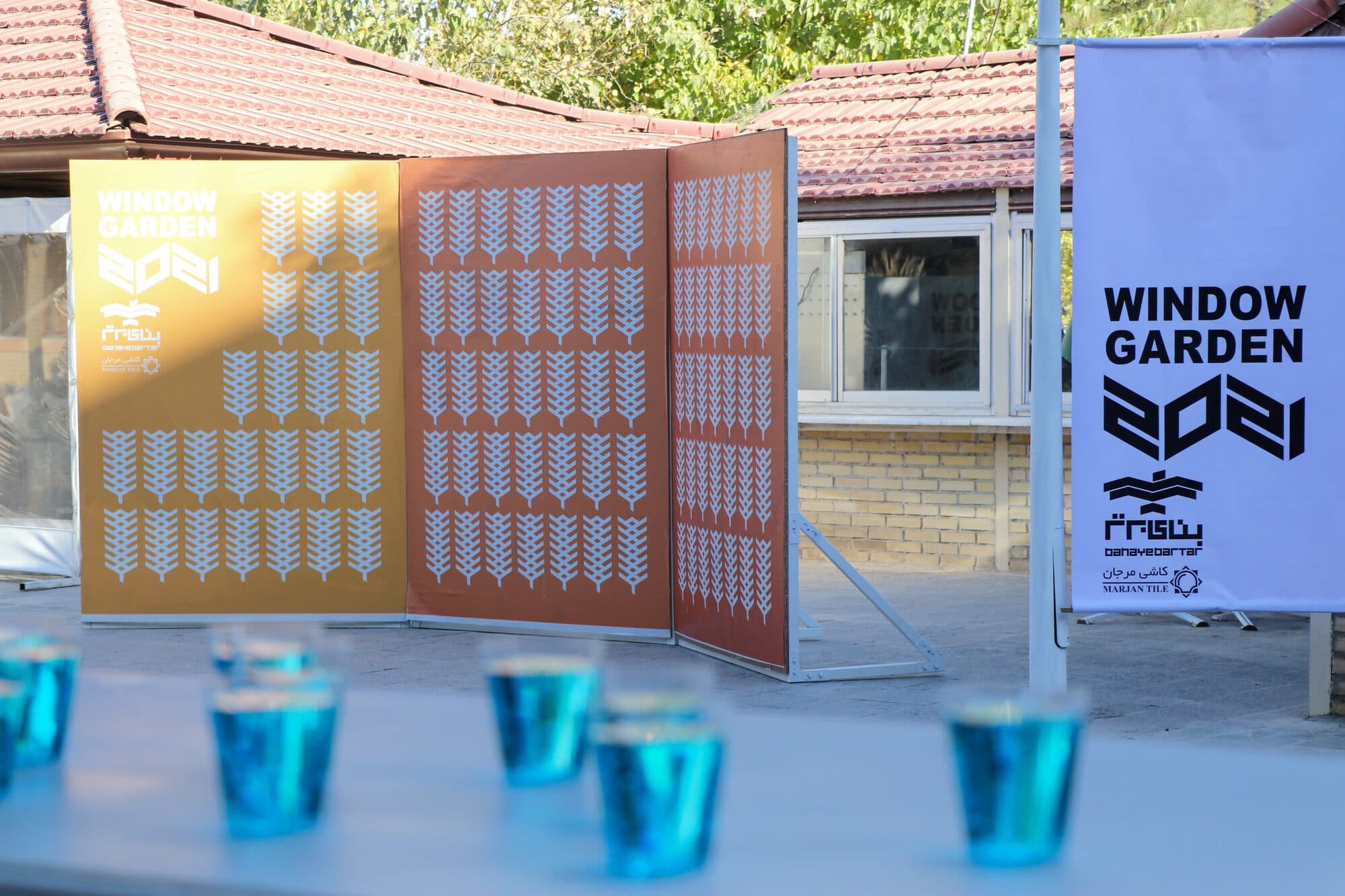 Perforated screens in ochre and rust standing open on the terrace, a WINDOW GARDEN 2021 banner beside them and cobalt glasses on the counter in front