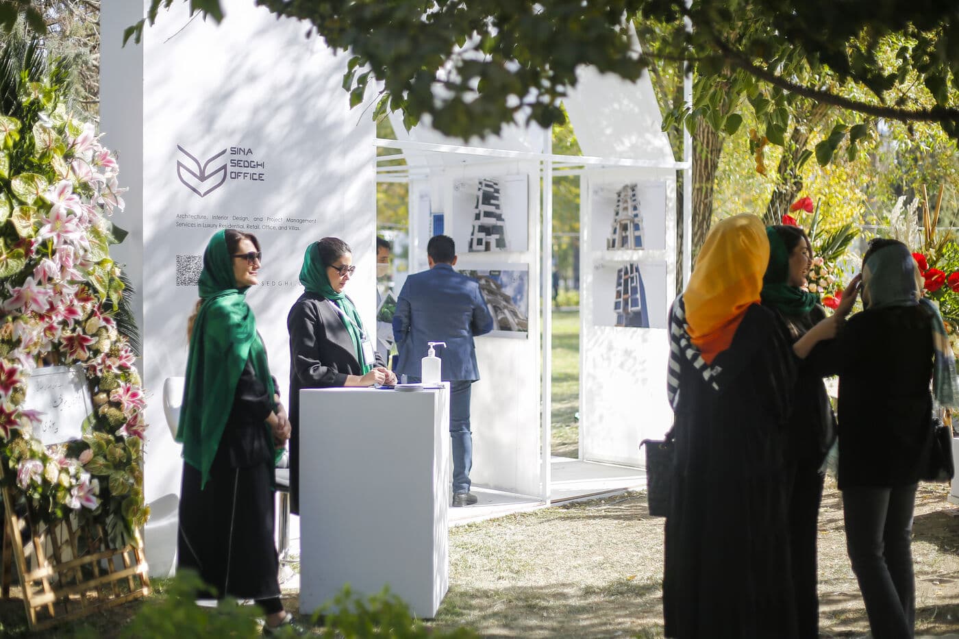 Visitors at a white pavilion in dappled light, mid-conversation with the exhibitor
