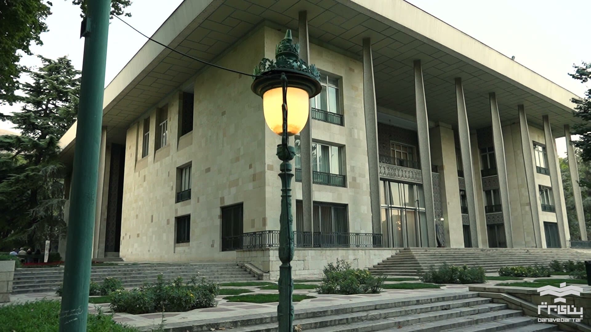The main pavilion of the Niavaran palace complex at dusk, its stone colonnade lit from below and a lamp standing on the path in front of it