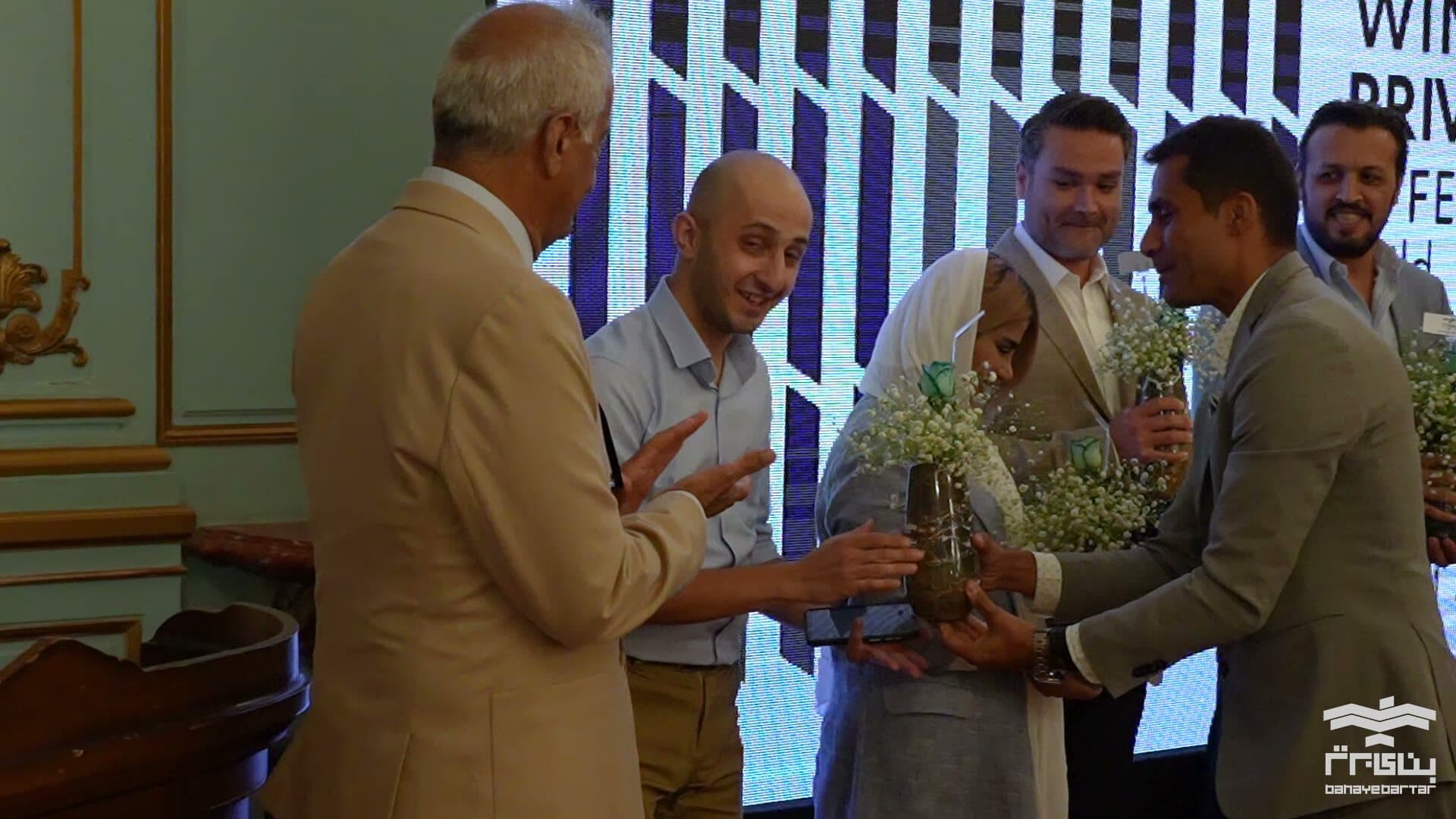 A gift passed between guests in one of the palace's gilded rooms, the WINDOW GARDEN private exhibition screen lit behind them