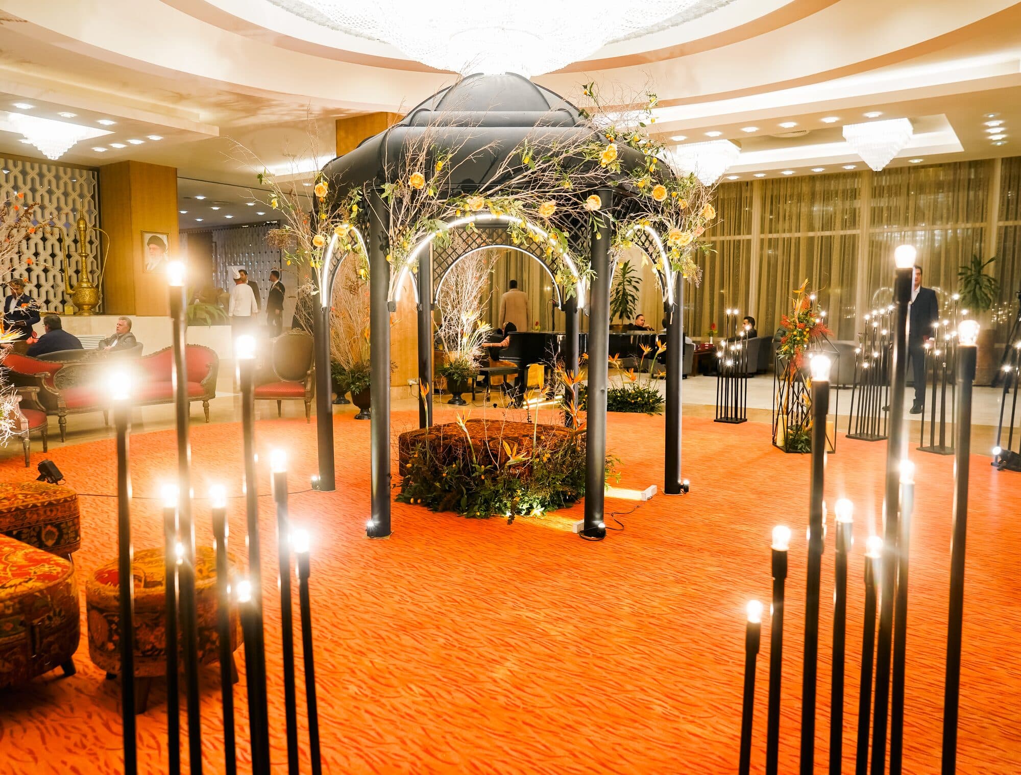 A pavilion of bare branches hung with pomegranates standing on a deep orange carpet in the hotel lobby, ringed by candle stands with armchairs set out around it