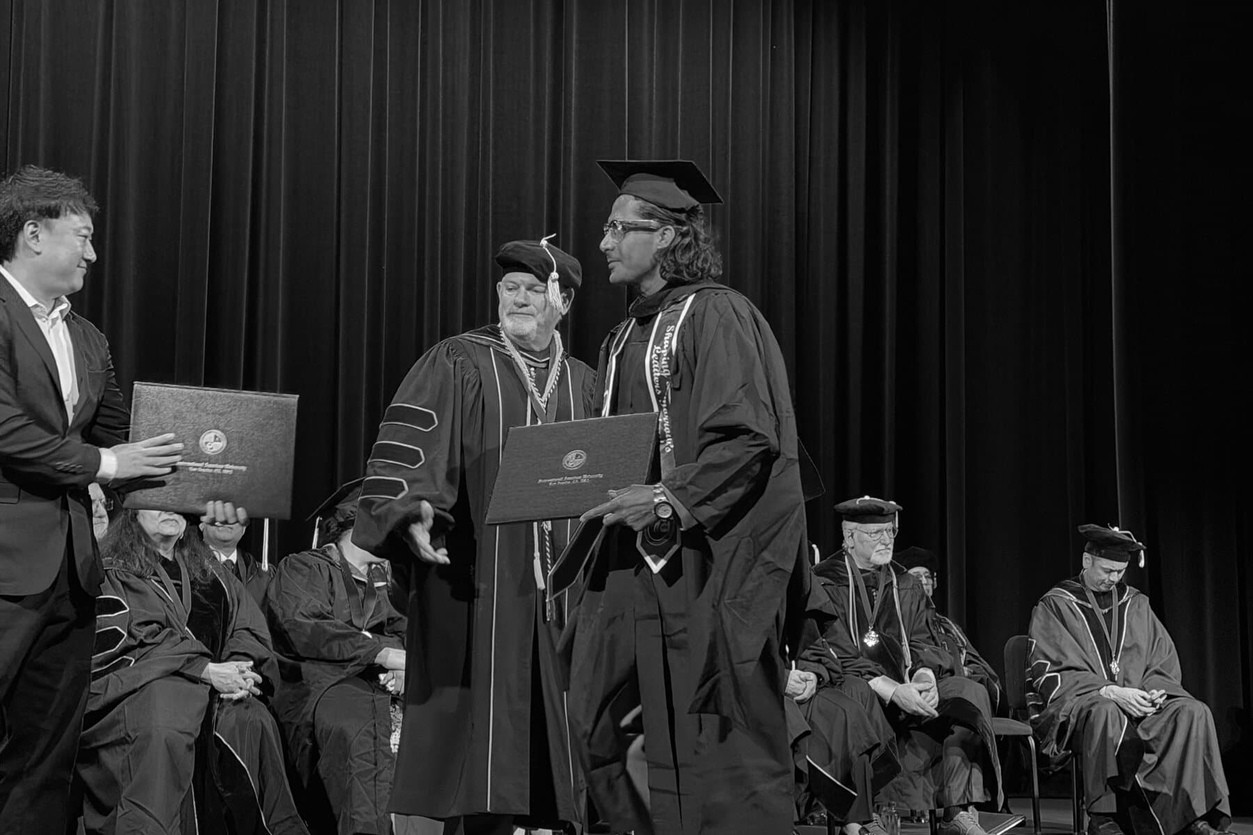 Ehsan Kaykhah crossing the stage in cap and gown, taking the diploma folder as the faculty row looks on