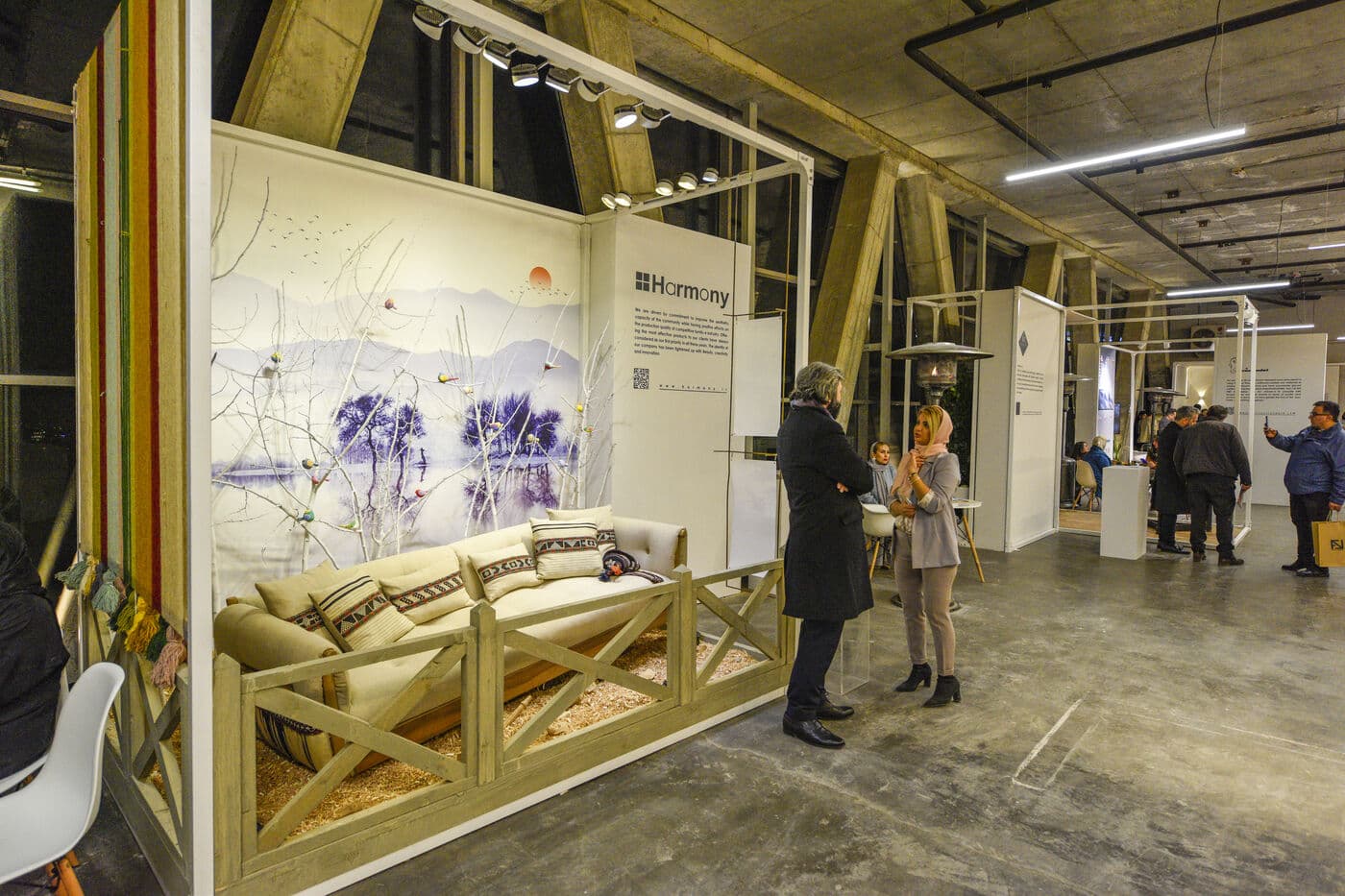 Visitors at a timber-framed living-room setting in the concrete hall — WINDOW DISPLAY, Tehran 2020