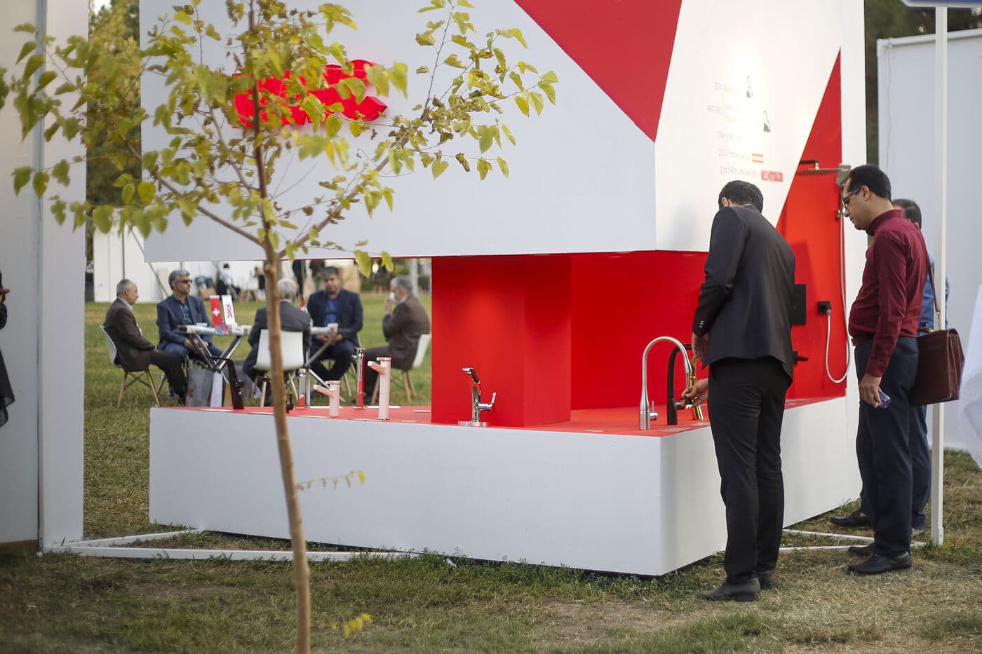 Two visitors at a red kitchen setting built around a tree in the garden — WINDOW GARDEN, Mashhad 2021