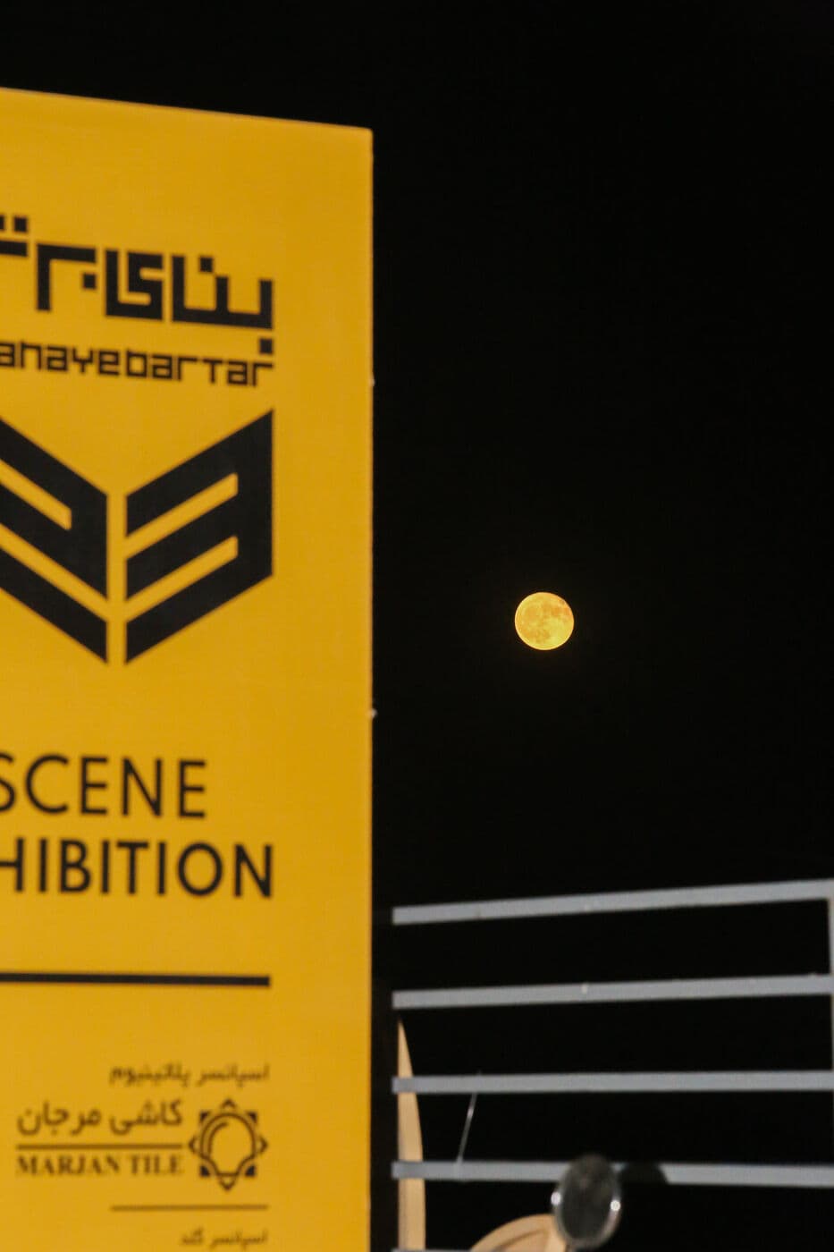 The yellow SCENE EXHIBITION sign under a full moon — WINDOW SCENE, Mashhad 2023